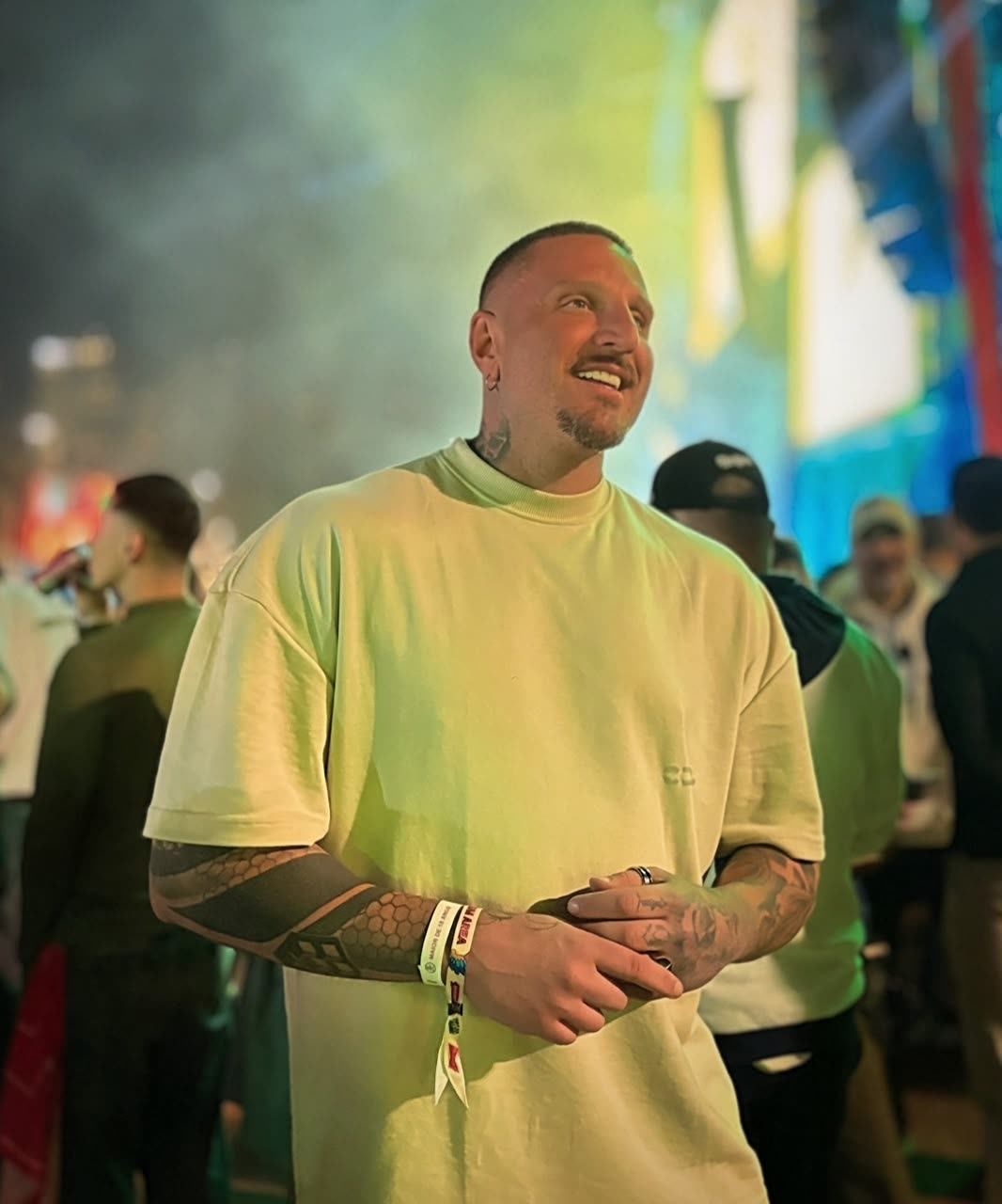 Guilherme Pallesi de camiseta clara, em pé no meio do público de um evento, com um palco iluminado ao fundo.